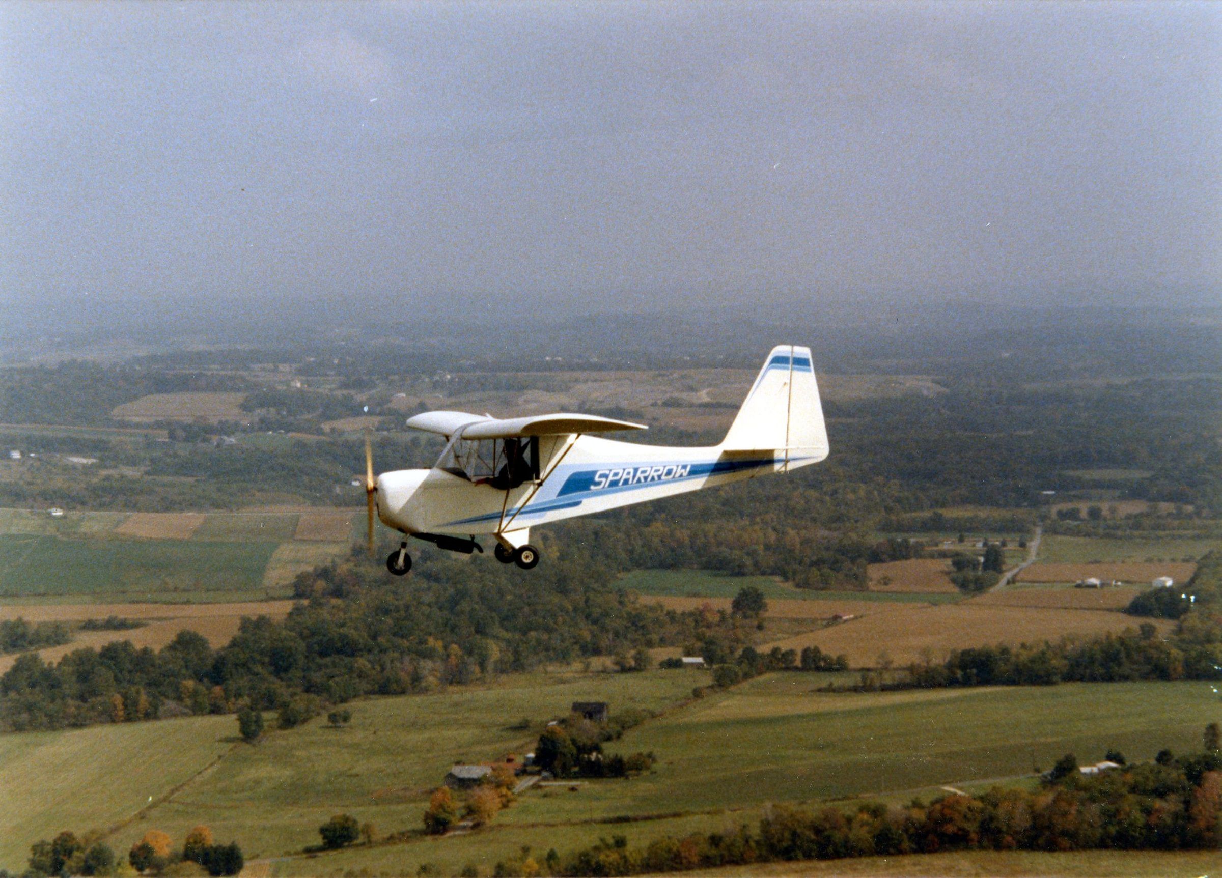 White and blue Carlson Sparrow in flight over farmland
