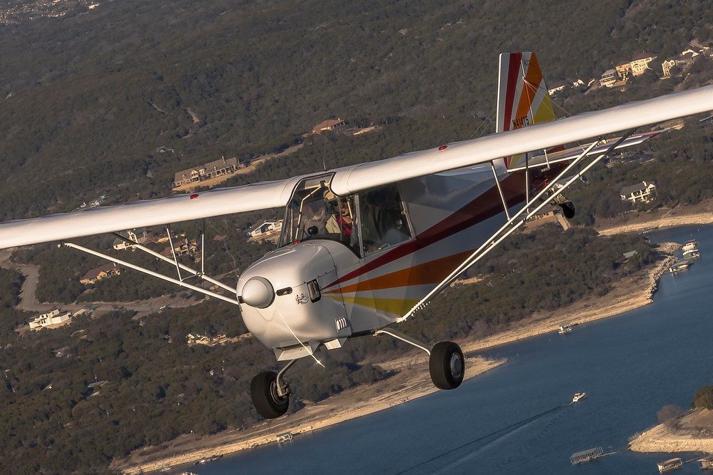 White Carlson Sparrow Sport Special with red, orange and yellow trim flying over a lake, tailwheel landing gear visible