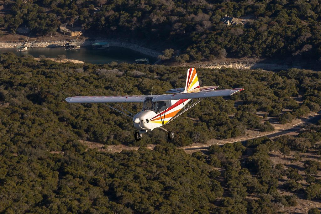 Carlson Sparrow Sport Special in flight over cedar-covered hills with a lake behind