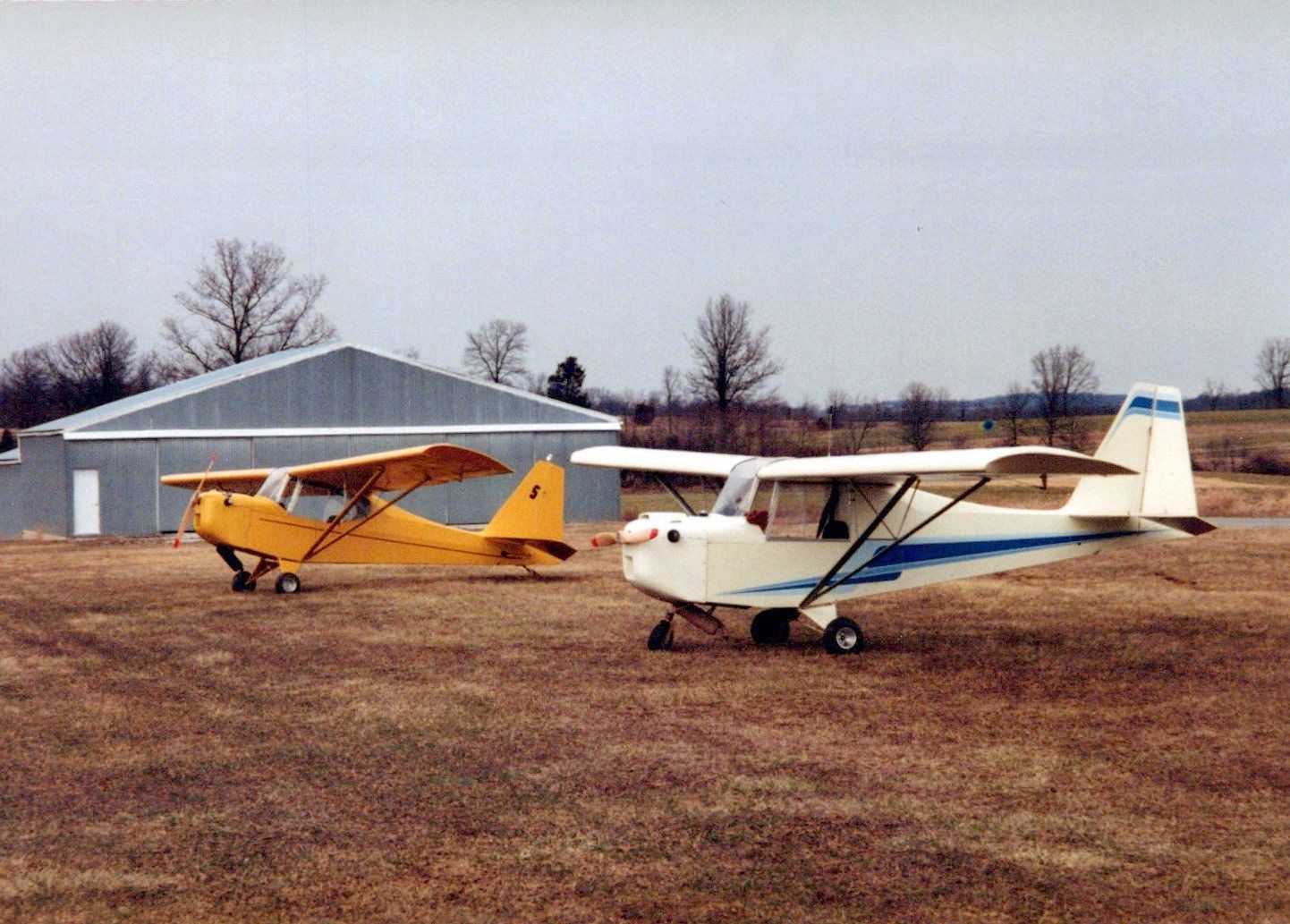A yellow and a white Carlson Sparrow parked side by side on a grass airfield