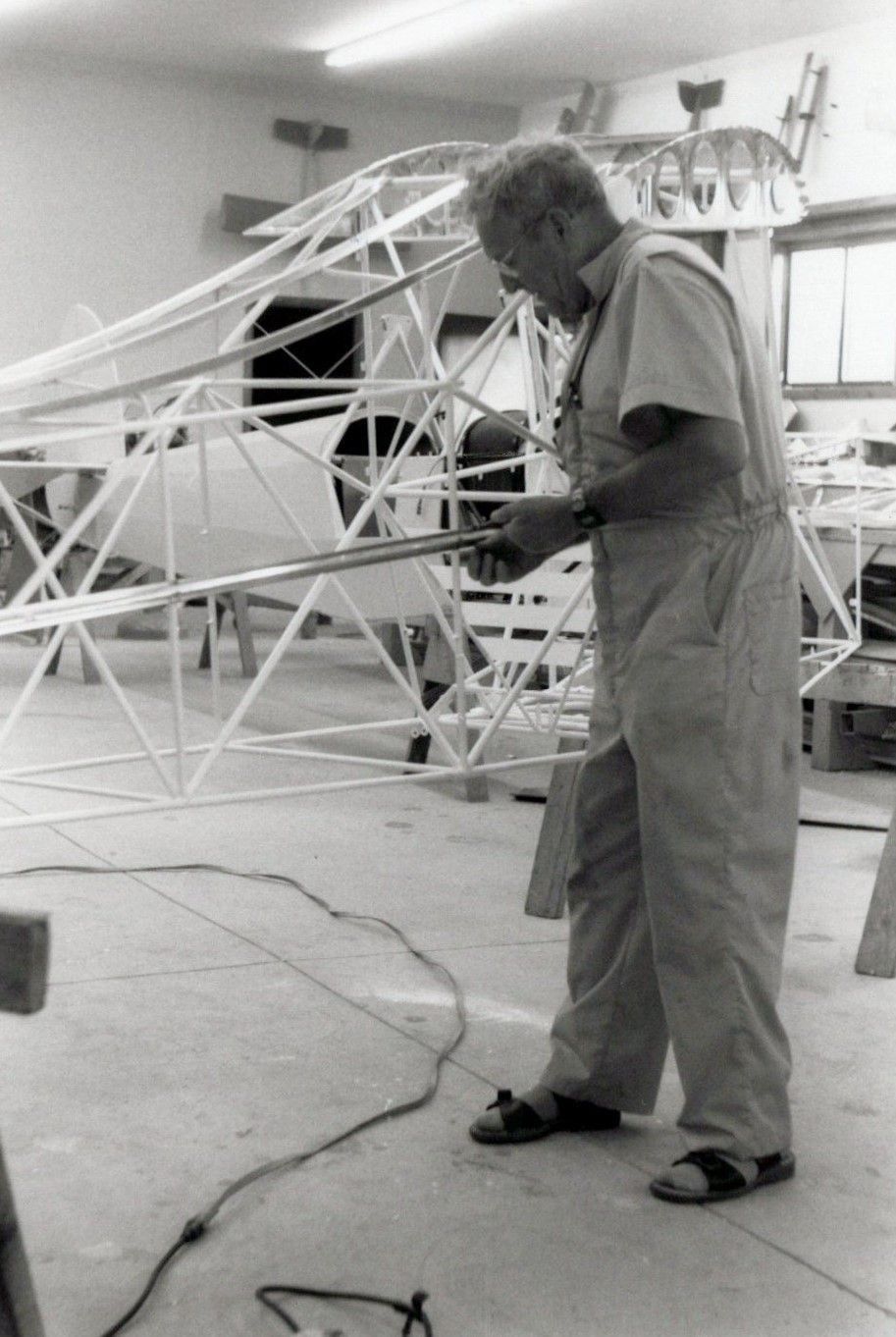 Black-and-white photograph of a man working on welded steel-tube fuselage frames in a shop